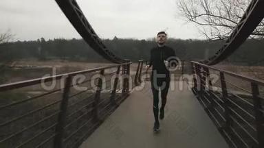 男子在桥上横渡河，前景。 白人年轻人穿着运动服在灰色阴天的公园里户外跑步。 什么